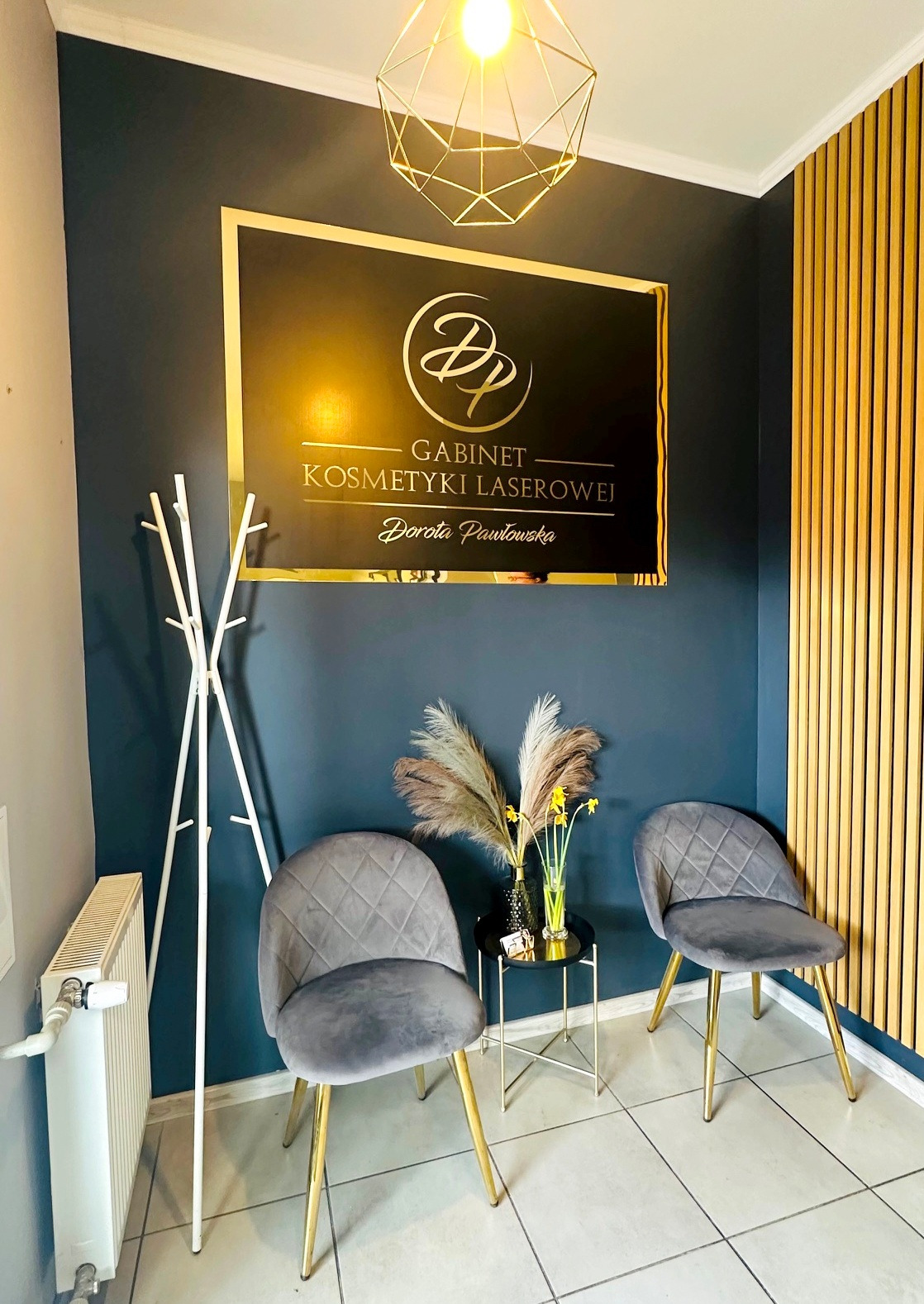 Poczekalnia gabinetu kosmetyki laserowej: dwie szare, welurowe krzesła, stolik z kwiatami, złote elementy dekoracyjne i logo na ciemnej ścianie.