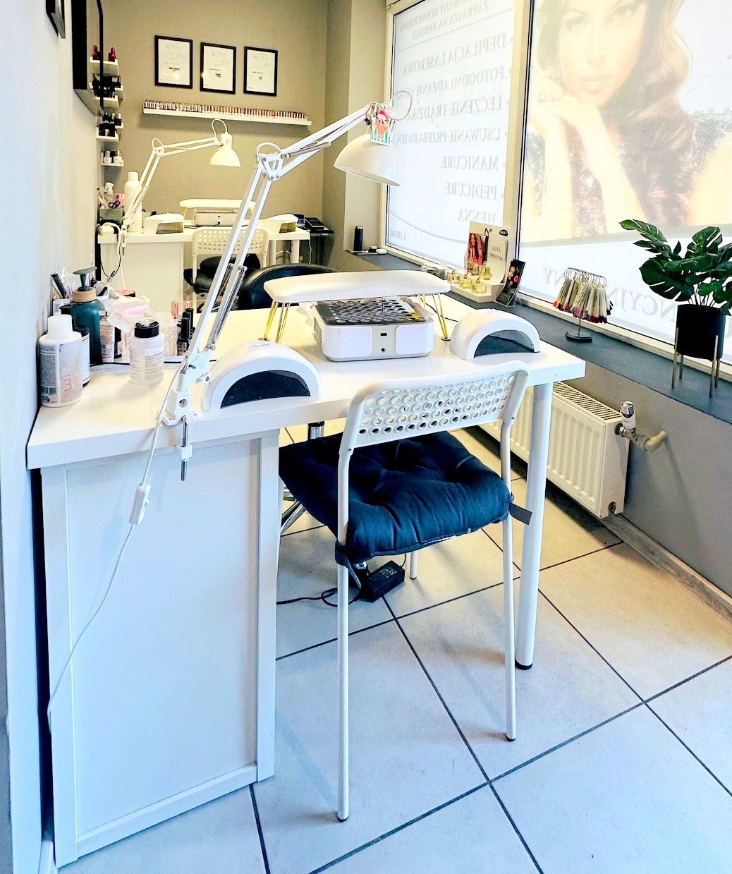 Jasne wnętrze salonu z białym stanowiskiem do manicure, lampą LED i krzesłem z poduszką. Na ścianie półki z lakierami i grafiki w ramkach.