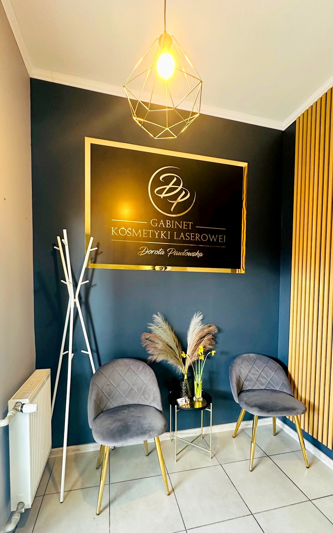 Eleganckie wnętrze: poczekalnia gabinetu kosmetyki laserowej z szarymi krzesłami, złotymi akcentami i designerską lampą. Na ścianie logo firmy.