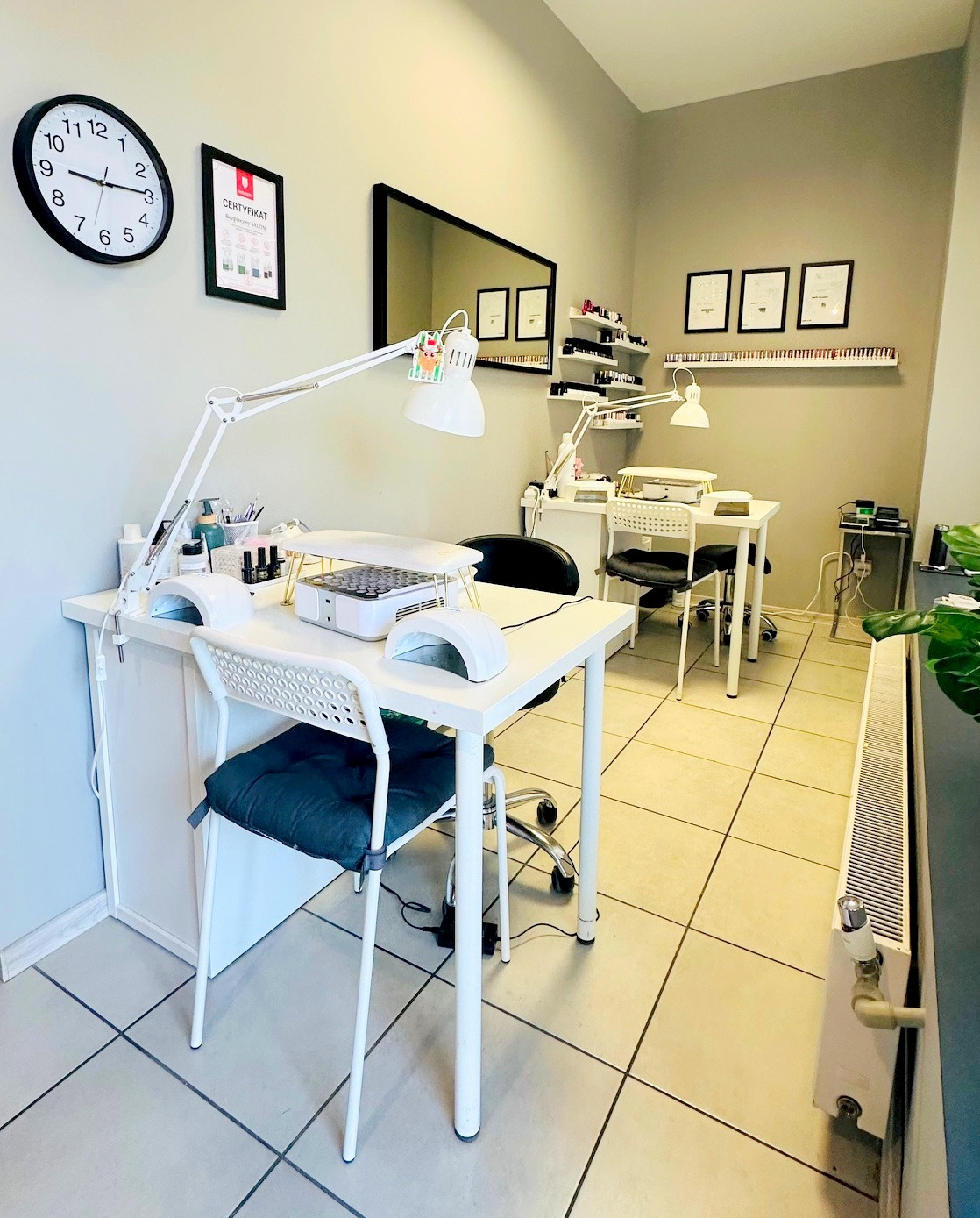 Jasne wnętrze salonu w Łodzi: stanowiska do manicure z lampami, certyfikat na ścianie i półki z lakierami. Minimalistyczny design, czyste i profesjonalne otoczenie.