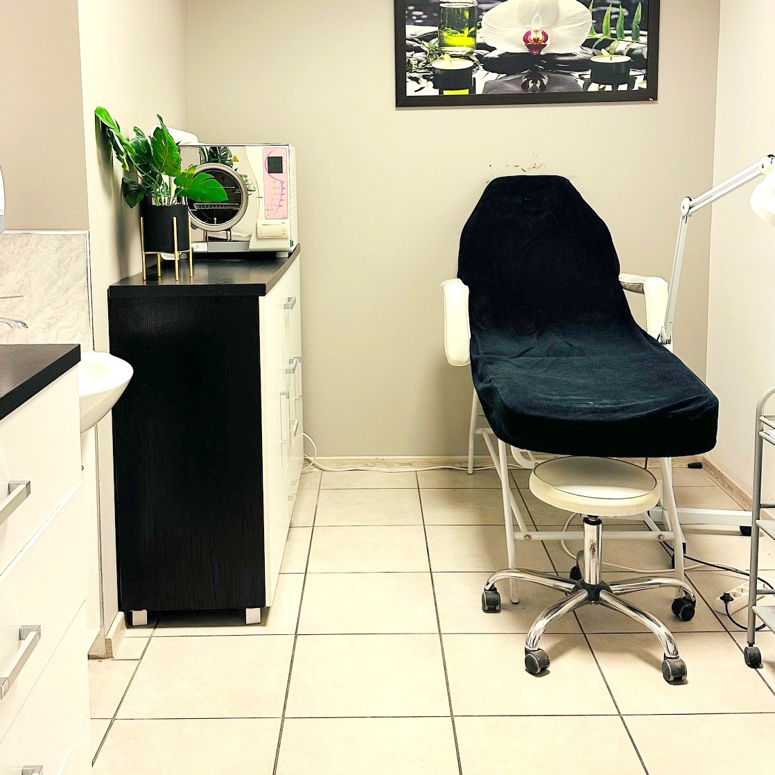 Jasne wnętrze gabinetu kosmetycznego z fotelem zabiegowym, sterylizatorem i rośliną w czarnej doniczce. Nad fotelem wisi obraz z motywem kwiatowym.