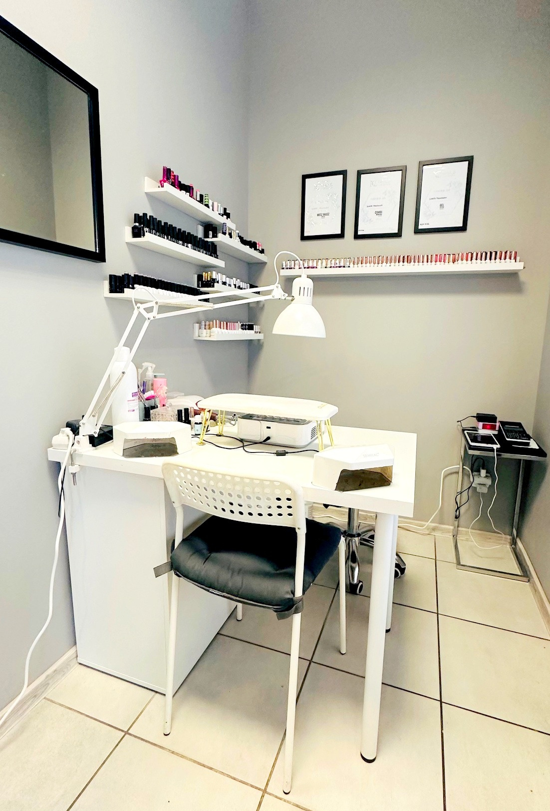 Stanowisko do manicure w salonie kosmetycznym: białe biurko z lampą, pochłaniacz pyłu, lakiery na półkach i certyfikaty na ścianie w minimalistycznym wnętrzu.