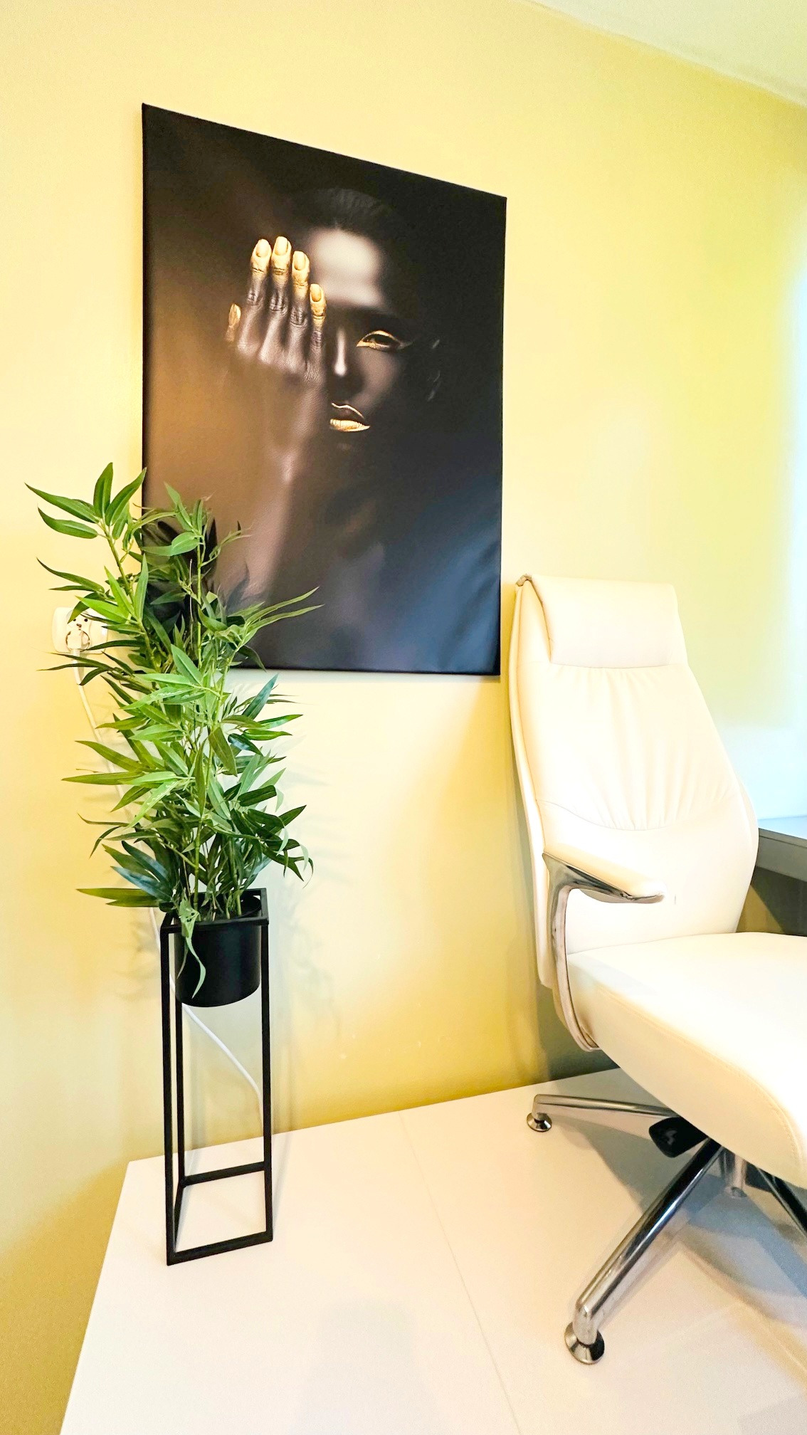 Stylowe wnętrze: biurko z rośliną w czarnej donicy i krzesło na tle ściany z obrazem kobiety z pomalowanymi złotem ustami i dłońmi.