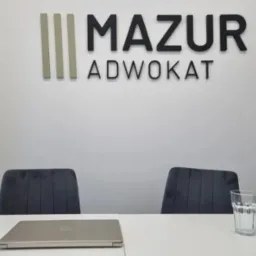 Biuro adwokackie: logo 'Mazur Adwokat' na ścianie, stół z laptopem i szklanką wody, dwa krzesła. Minimalistyczne wnętrze, gotowe na spotkanie z klientem.