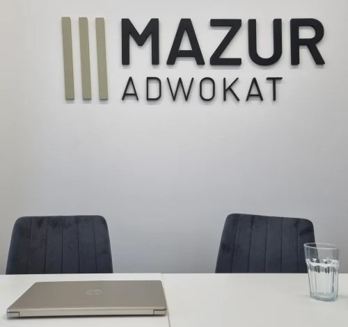 Biuro adwokackie: logo 'Mazur Adwokat' na ścianie, stół z laptopem i szklanką wody, dwa krzesła. Minimalistyczne wnętrze, gotowe na spotkanie z klientem.