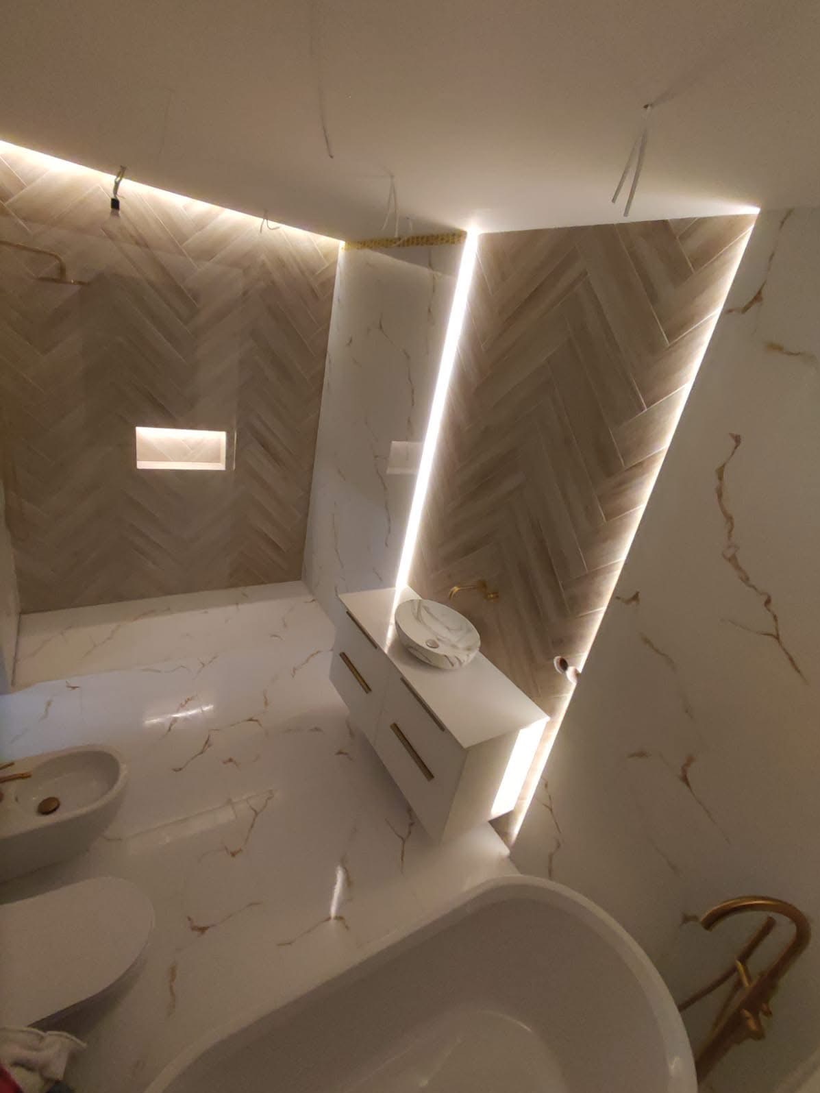 Elegancka łazienka z marmurową podłogą i ścianami, oświetleniem LED, białą szafką z umywalką i złotą armaturą. Widoczna wanna i bidet.