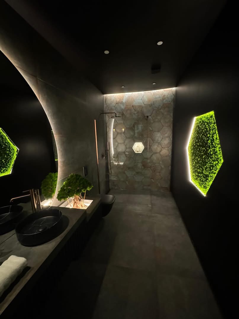 Nowoczesna łazienka w ciemnej tonacji z heksagonalnymi płytkami pod prysznicem, podświetlanymi panelami z mchu i umywalką z rośliną bonsai.