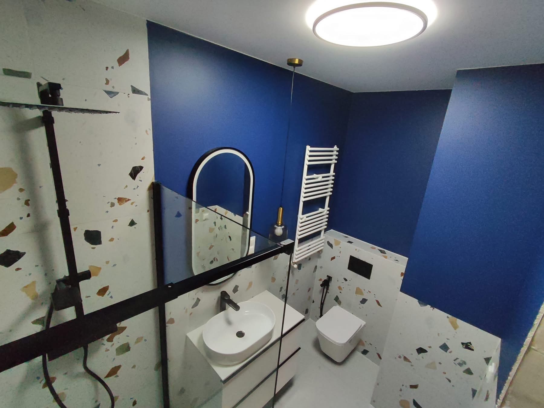 Niebiesko-biała łazienka z kabiną typu walk-in, umywalką nablatową i grzejnikiem drabinkowym. Ściany zdobią płytki lastryko i głęboki odcień niebieskiego.