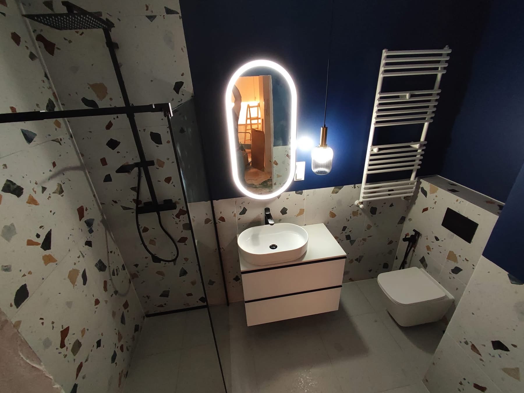 Nowoczesna łazienka z umywalką na szafce, podświetlanym lustrem i grzejnikiem drabinkowym na tle granatowej ściany i płytek lastryko.