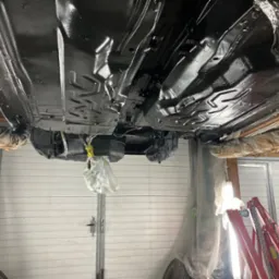 Podwozie auta po konserwacji antykorozyjnej, widoczne z dołu na podnośniku. Elementy zawieszenia, ramy i karoserii pokryte czarną, błyszczącą powłoką ochronną.