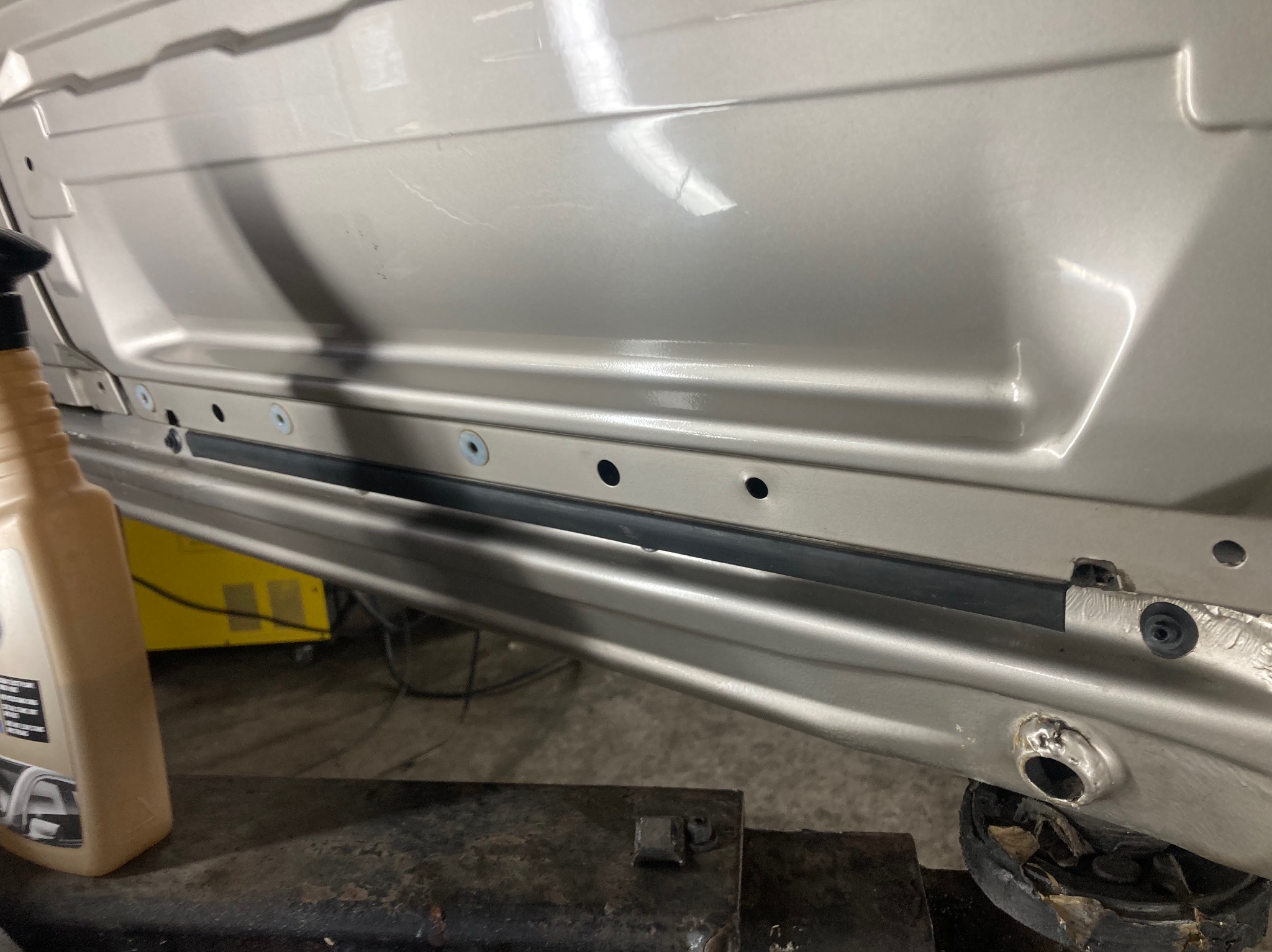 Fragment karoserii auta w warsztacie, widoczne otwory montażowe, uszczelka i butelka środka konserwującego. Element poddawany naprawie blacharskiej.