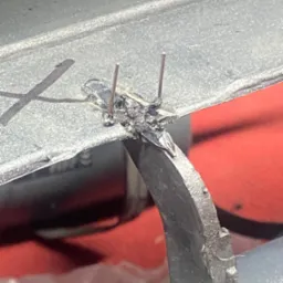 Spoina łącząca dwa elementy metalowe, widoczne ślady spawania i dwa cienkie pręty wystające ze spoiny. Detal metalowej konstrukcji na czerwonym tle.