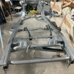 Szara, odnowiona rama samochodowa po piaskowaniu i malowaniu, widoczna w warsztacie z elementami wyposażenia w tle. Metalowa konstrukcja gotowa do montażu.