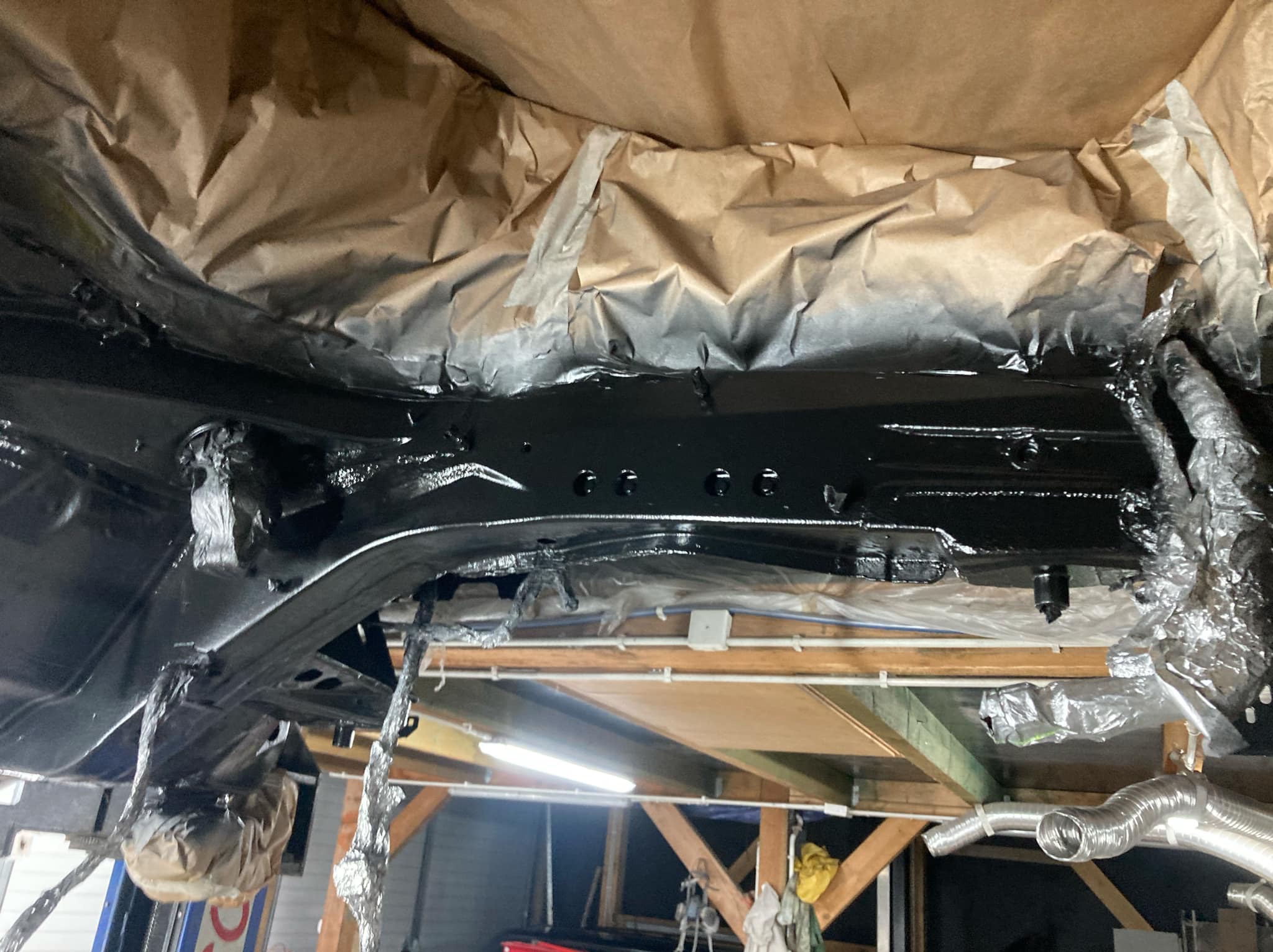 Podwozie samochodu po nałożeniu czarnej powłoki antykorozyjnej, zabezpieczone folią i papierem. Widoczne elementy konstrukcyjne pojazdu na podnośniku.