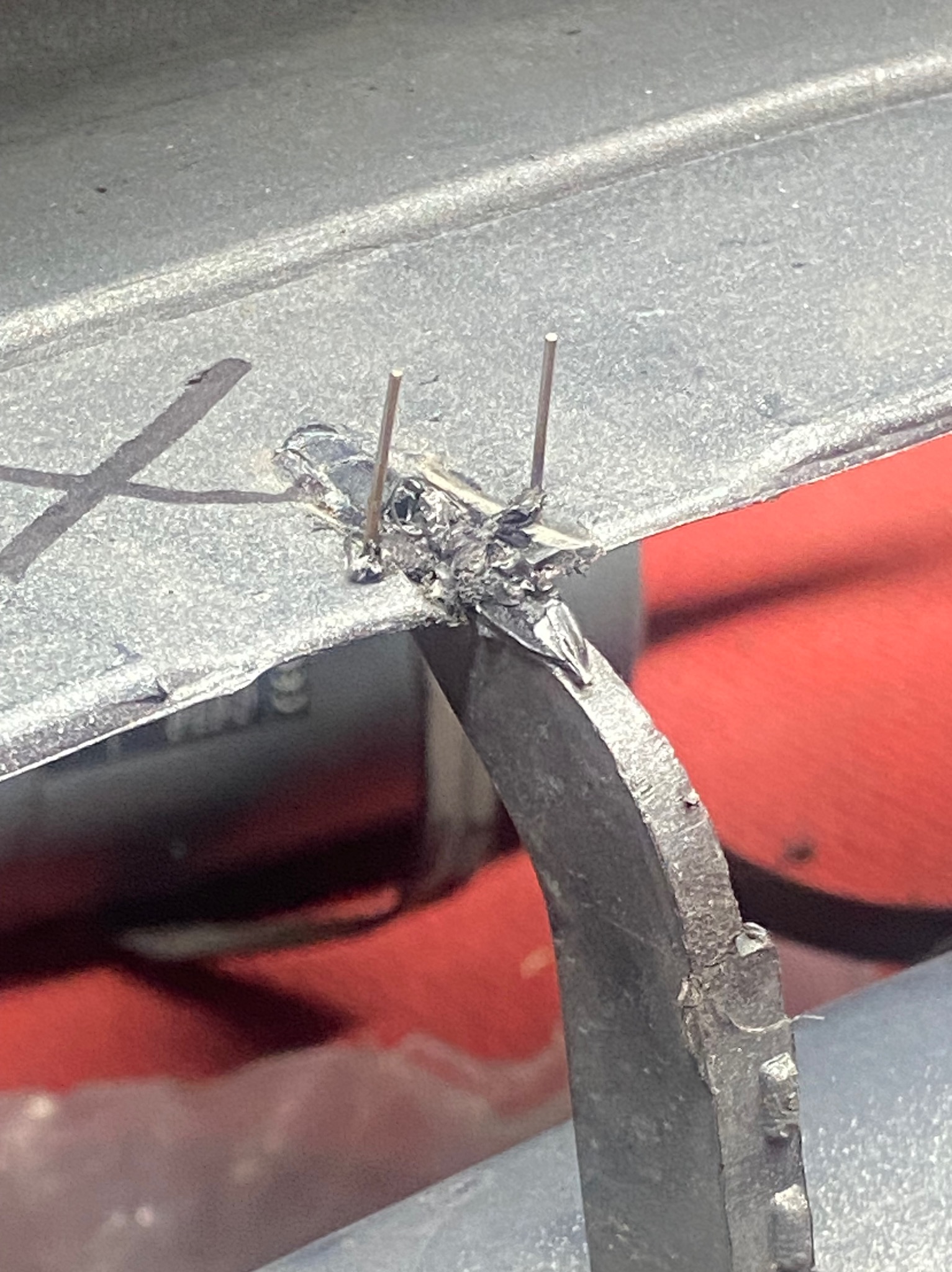 Spoina łącząca dwa elementy metalowe, widoczne ślady spawania i dwa cienkie pręty wystające ze spoiny. Detal metalowej konstrukcji na czerwonym tle.