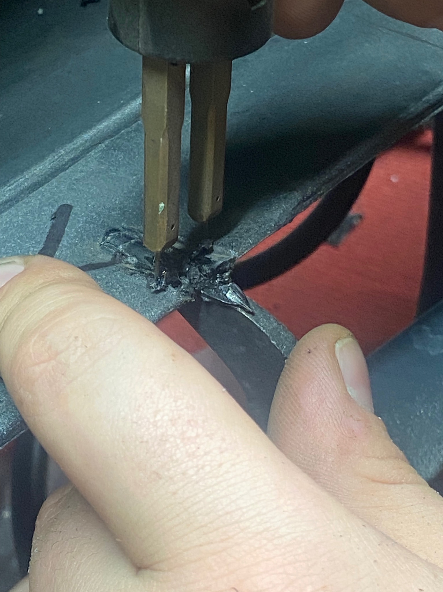 Zbliżenie na proces spawania plastiku z użyciem spawarki, widoczne dłonie operatora i stopiony plastik na szarej powierzchni, z czerwonym elementem w tle.