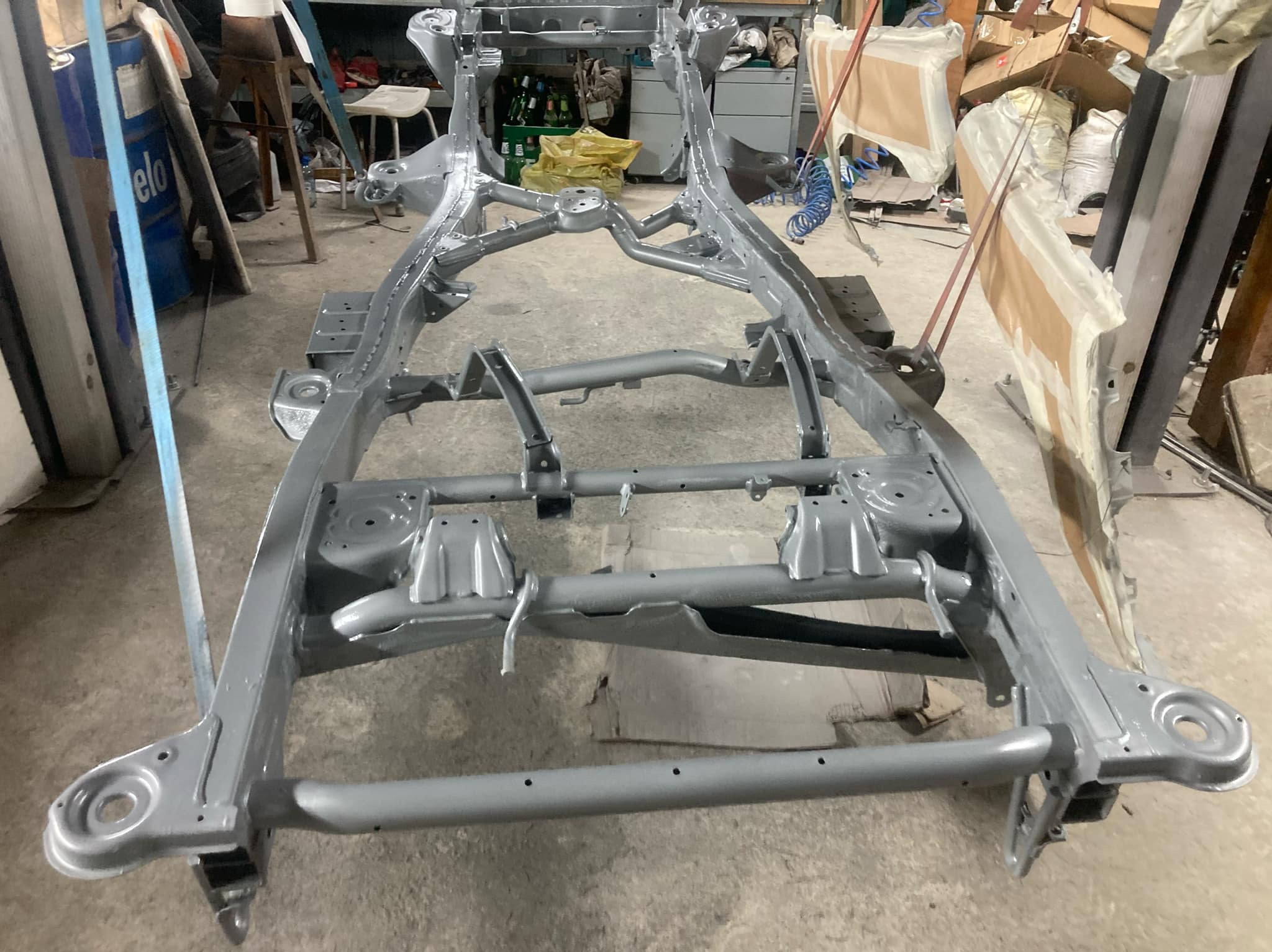 Szara, odnowiona rama samochodowa po piaskowaniu i malowaniu, widoczna w warsztacie z elementami wyposażenia w tle. Metalowa konstrukcja gotowa do montażu.