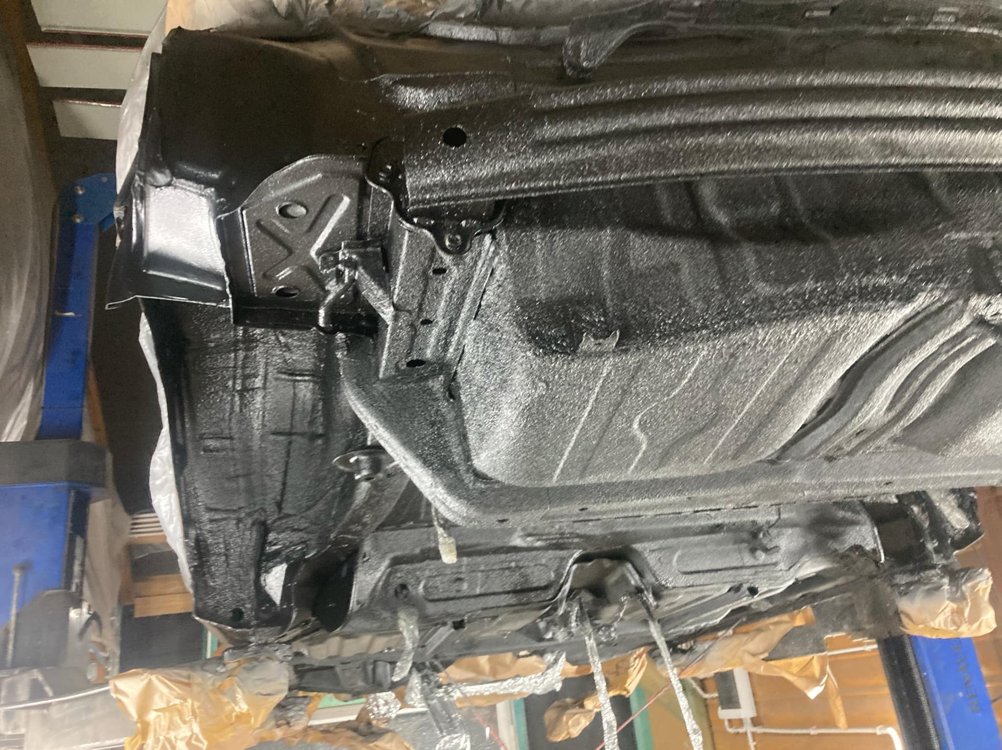 Podwozie auta po konserwacji antykorozyjnej, zabezpieczone czarną powłoką. Widoczny podnośnik warsztatowy i elementy zabezpieczone folią. Detal z bliska.