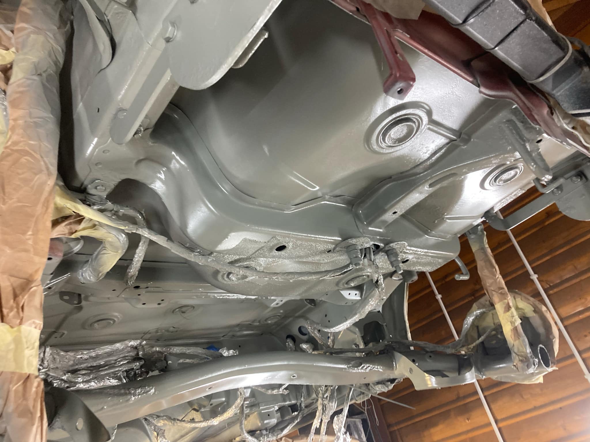 Podwozie auta po konserwacji, zabezpieczone przed korozją. Elementy zawieszenia i blachy pokryte szarą farbą, niektóre części zamaskowane folią i taśmą malarską.