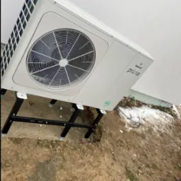 Biała jednostka zewnętrzna pompy ciepła Galmet Prima na czarnym stelażu, ustawiona na ziemi z resztkami śniegu. Widok z boku, blisko ściany budynku.