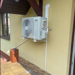 „Cimek” Michał Cimek - Zamontowana jednostka zewnętrzna klimatyzacji AUX DC Inverter na elewacji budynku z beżowym tynkiem, widoczne przewody i rury instalacyjne.