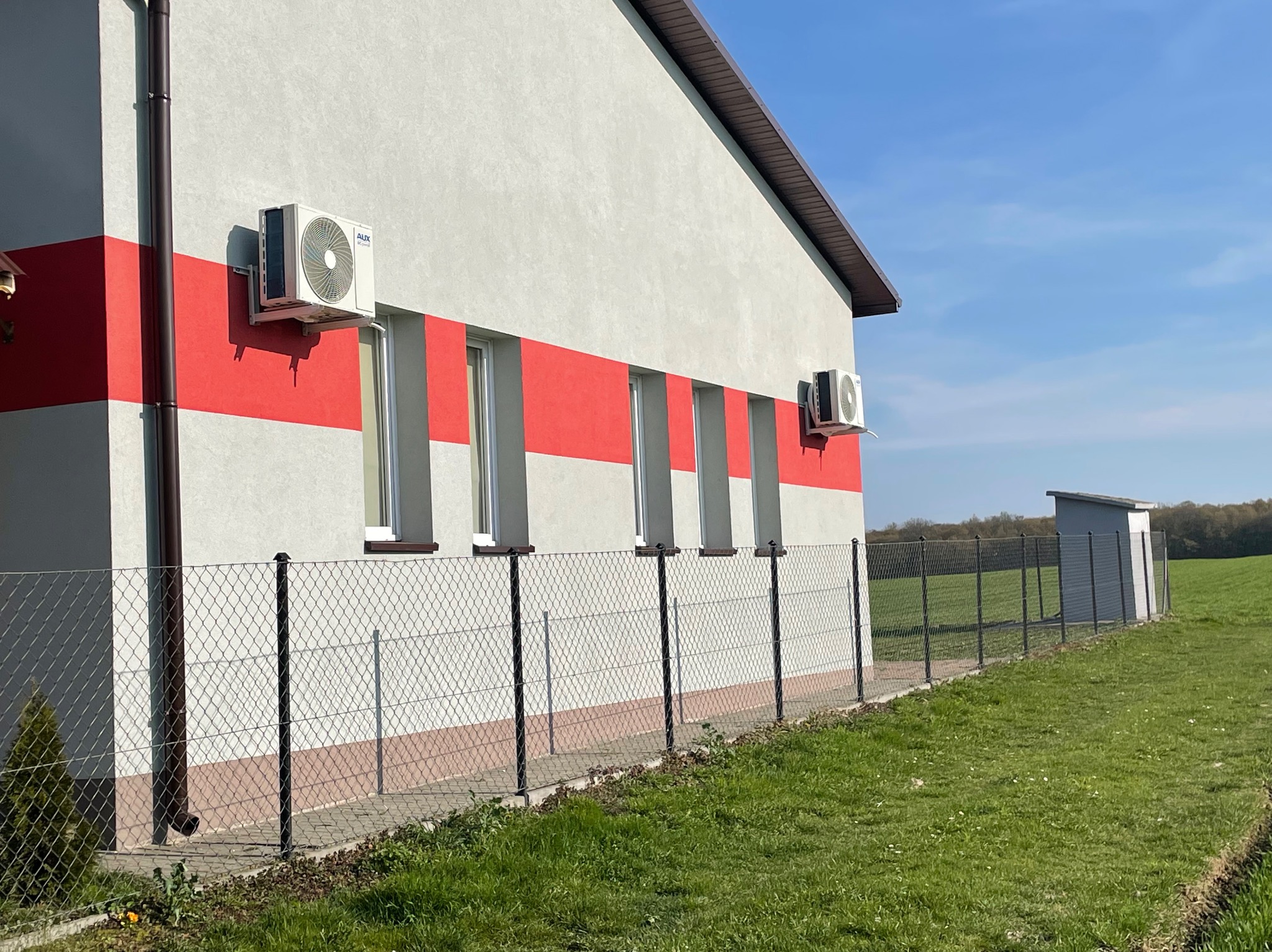 Biały budynek z dwoma zamontowanymi klimatyzatorami AUX, oddzielony ogrodzeniem z siatki od zielonego trawnika, w tle błękitne niebo.
