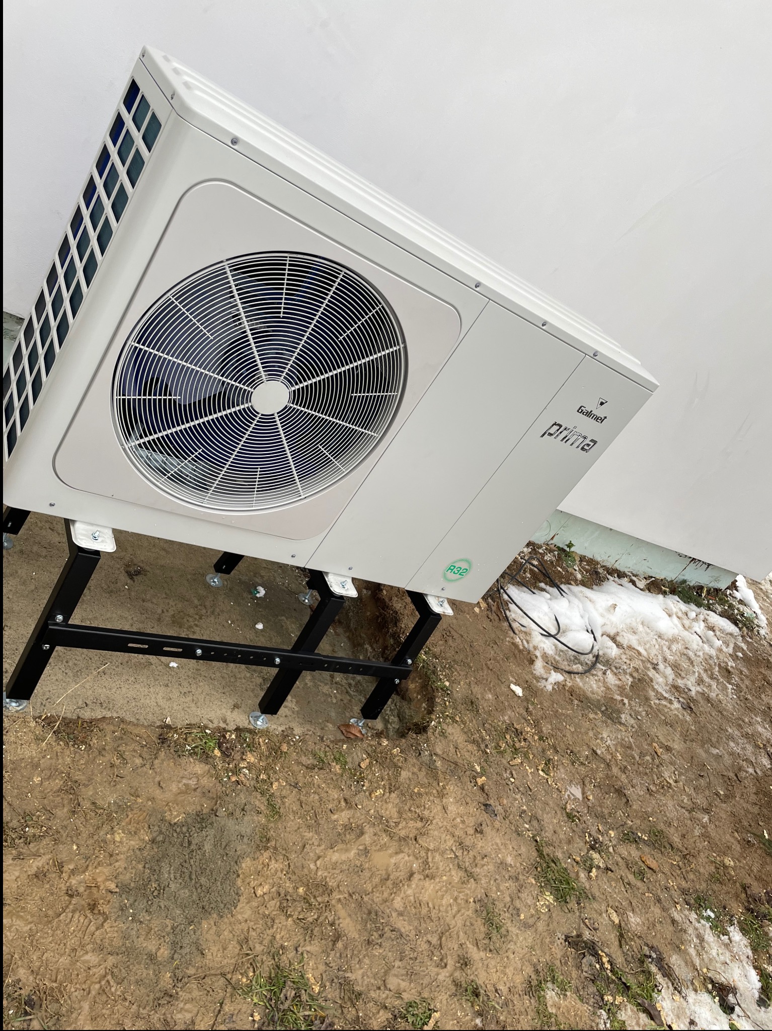 Biała jednostka zewnętrzna pompy ciepła Galmet Prima na czarnym stelażu, ustawiona na ziemi z resztkami śniegu. Widok z boku, blisko ściany budynku.