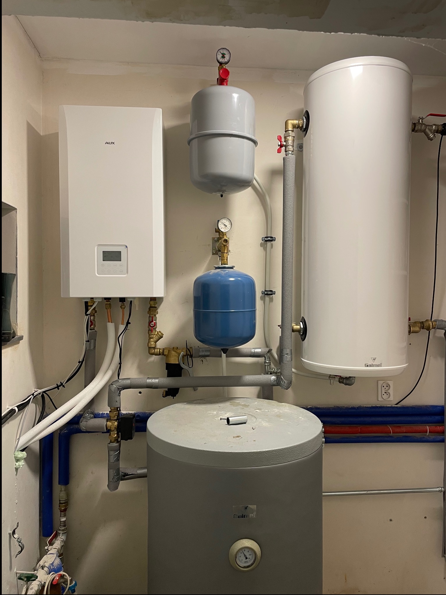 Kompletna instalacja grzewcza z białym kotłem AUX, szarym i białym zasobnikiem Galmet oraz niebieskim zbiornikiem wyrównawczym, widoczne rury i zawory.