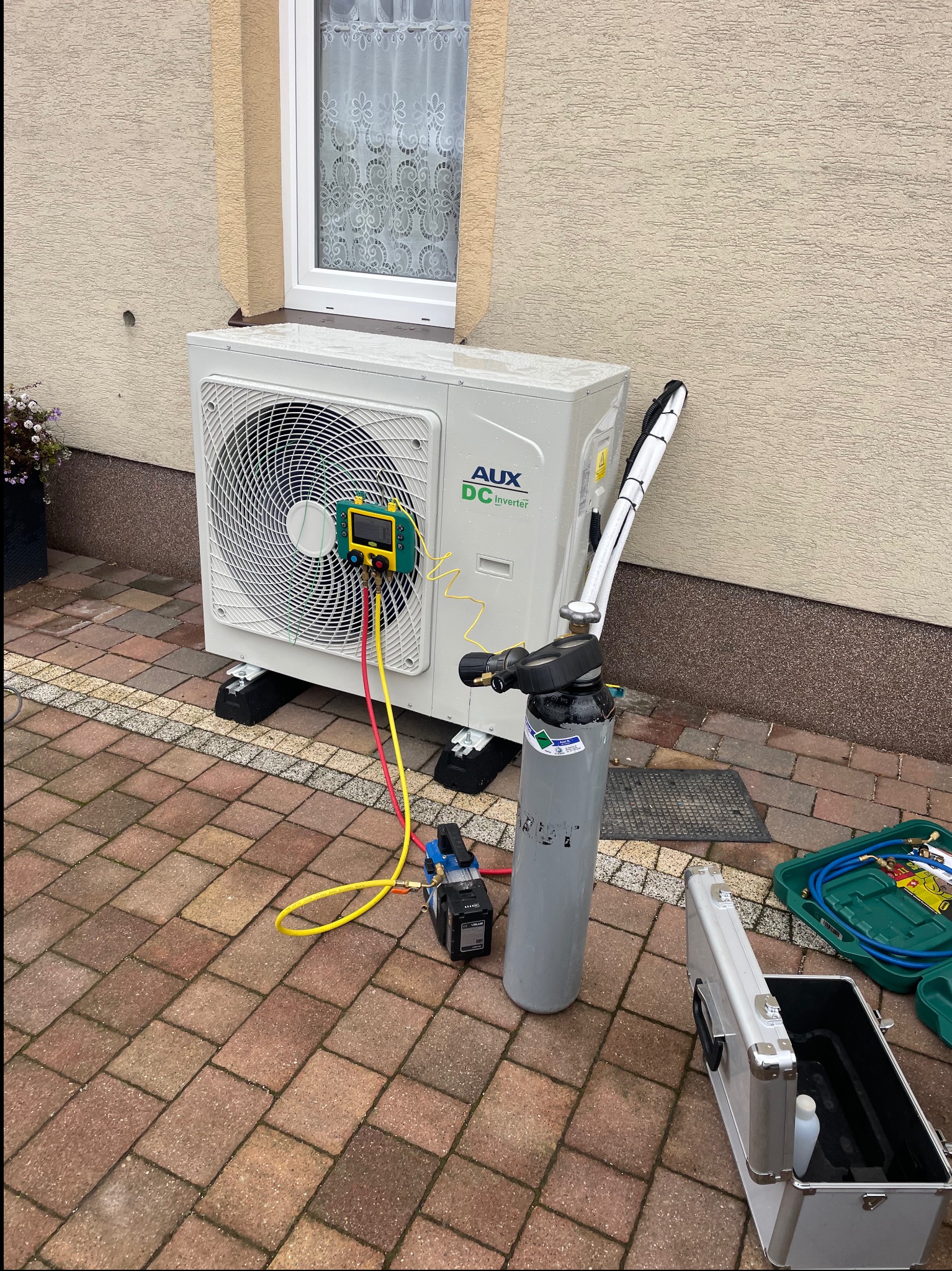 Biała jednostka zewnętrzna klimatyzacji AUX DC Inverter pod oknem, podłączona do manometru i butli z czynnikiem chłodniczym, na tle elewacji i kostki brukowej.