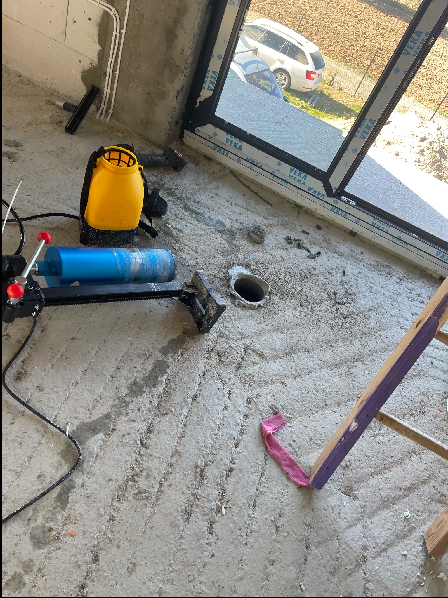 Wnętrze w trakcie prac instalacyjnych: wiertnica diamentowa z niebieskim wiertłem, żółty zbiornik, widoczne ślady wiercenia w posadzce, okno VEKA w tle.