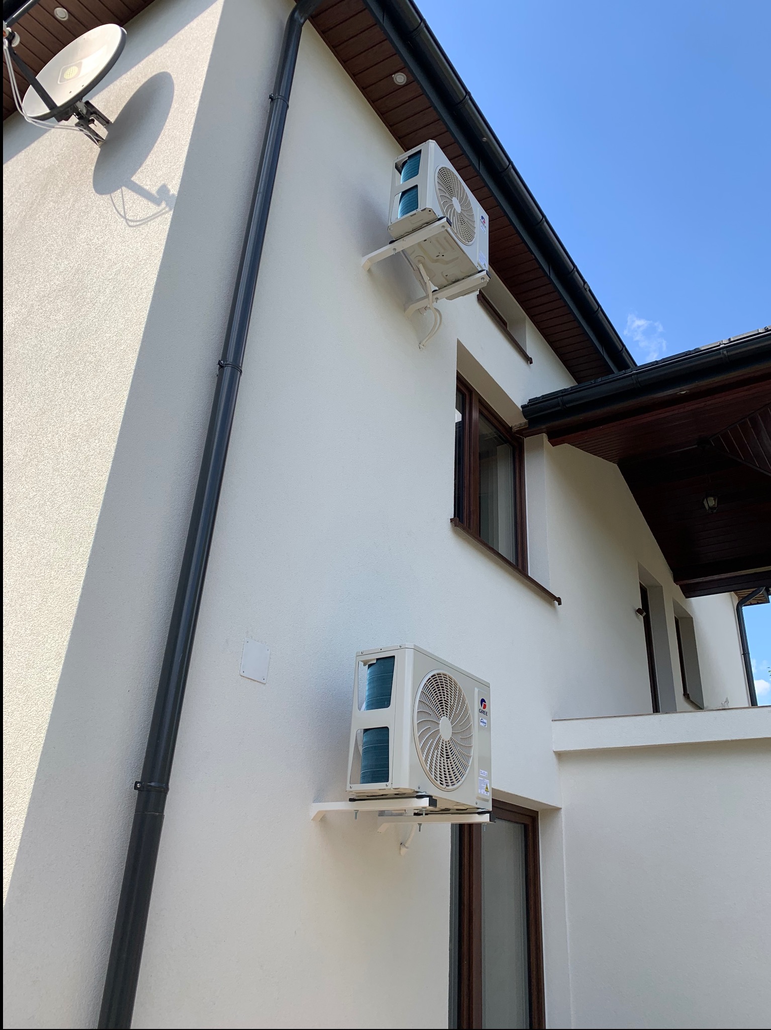 Dwa klimatyzatory zamontowane na elewacji domu w jasnej kolorystyce. Widoczne rynny, okna z brązowymi ramami i antena satelitarna. Błękitne niebo w tle.