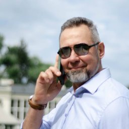 BNT SPÓŁKA Z OGRANICZONĄ ODPOWIEDZIALNOŚCIĄ - Mężczyzna w okularach przeciwsłonecznych rozmawia przez telefon, ubrany w błękitną koszulę, z budynkiem w tle. Widoczne niebo i zieleń.