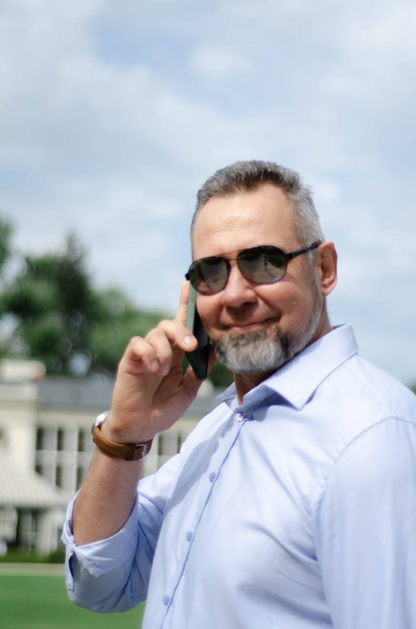 Mężczyzna w okularach przeciwsłonecznych rozmawia przez telefon, ubrany w błękitną koszulę, z budynkiem w tle. Widoczne niebo i zieleń.