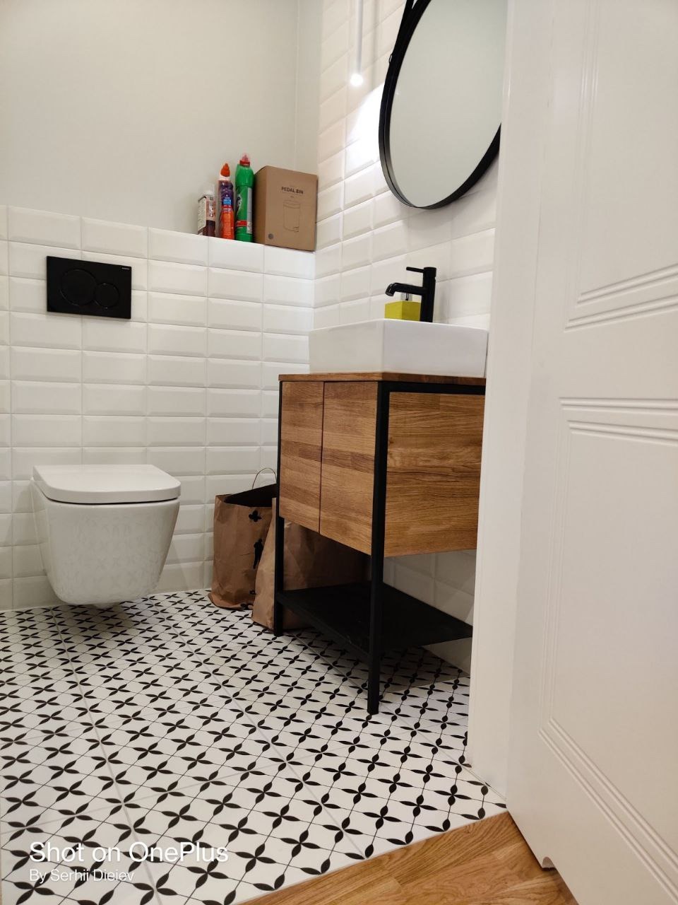 Nowoczesna łazienka po remoncie: białe płytki, wisząca toaleta, drewniana szafka z umywalką i czarne dodatki. Na podłodze wzorzyste płytki.