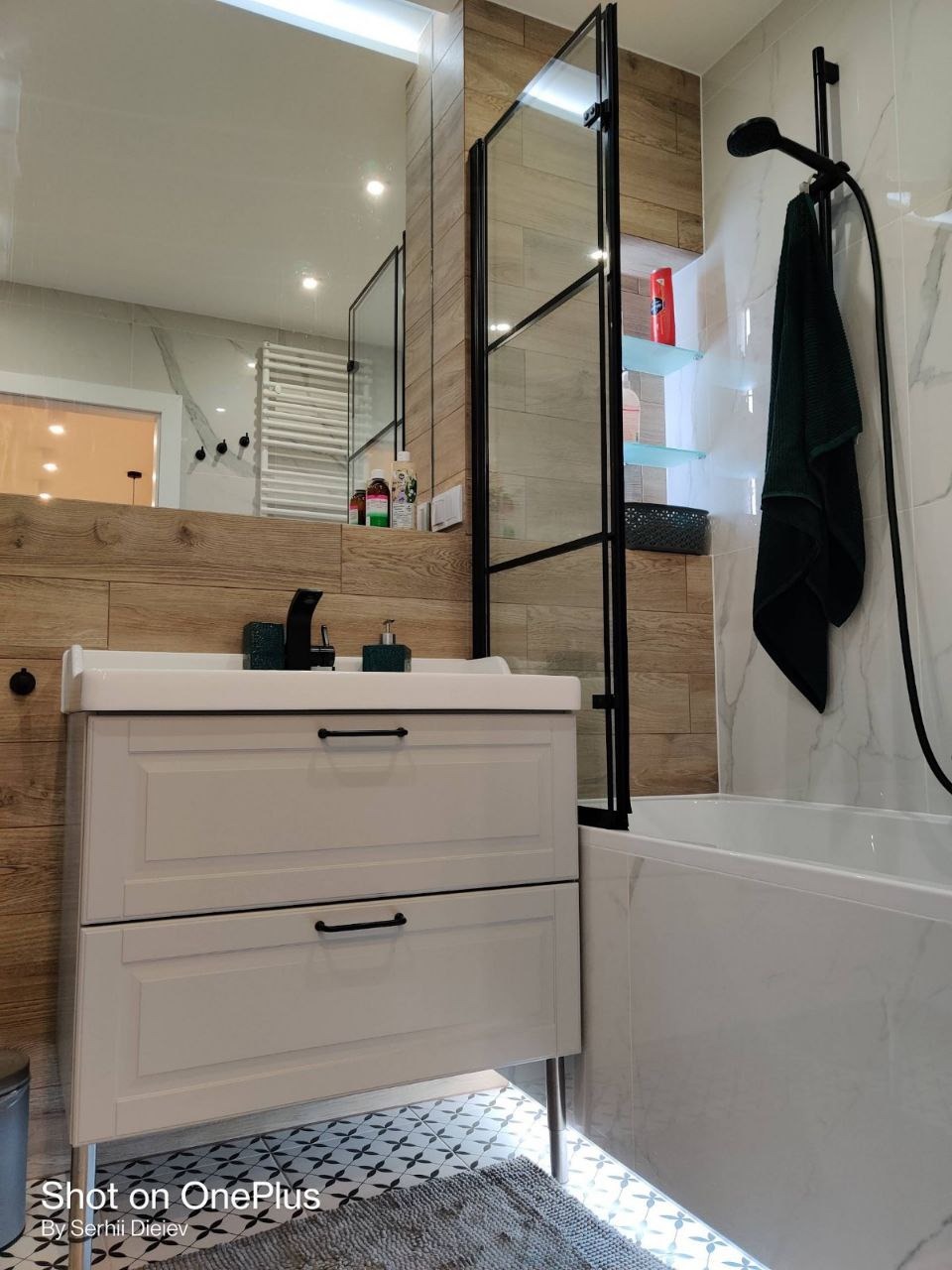 Nowoczesna łazienka z białą szafką pod umywalką, czarną armaturą i kabiną prysznicową w stylu loft, wykończona płytkami imitującymi drewno i marmur.