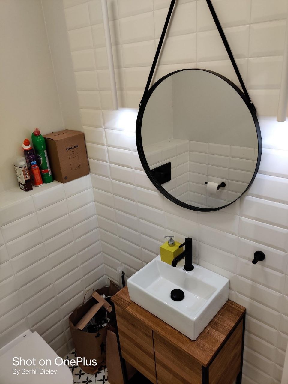 Biała umywalka nablatowa z czarną armaturą na drewnianej szafce w wykończonej łazience z białymi płytkami w stylu cegiełki i okrągłym lustrem.