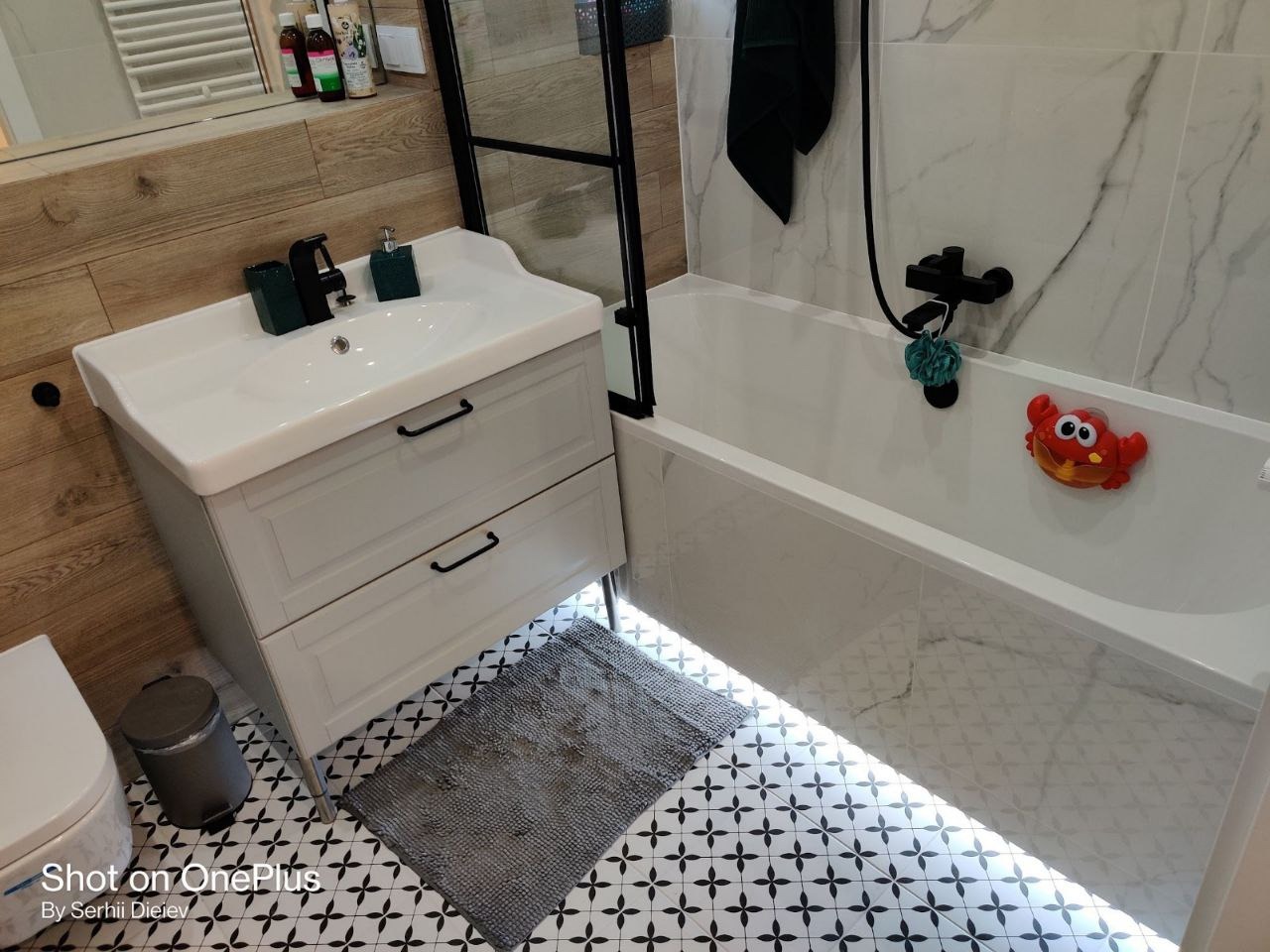 Nowoczesna łazienka z białą armaturą, drewnopodobnymi panelami ściennymi i wzorzystą podłogą. Uwagę przykuwa zabawka kraba na wannie.