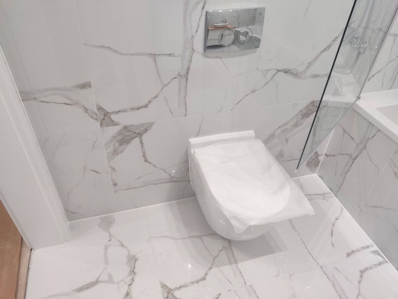 Biała toaleta podtynkowa z marmurowymi płytkami w wykończonej łazience. Nowoczesne rozwiązanie z przyciskiem spłukującym na ścianie i kabiną prysznicową w tle.