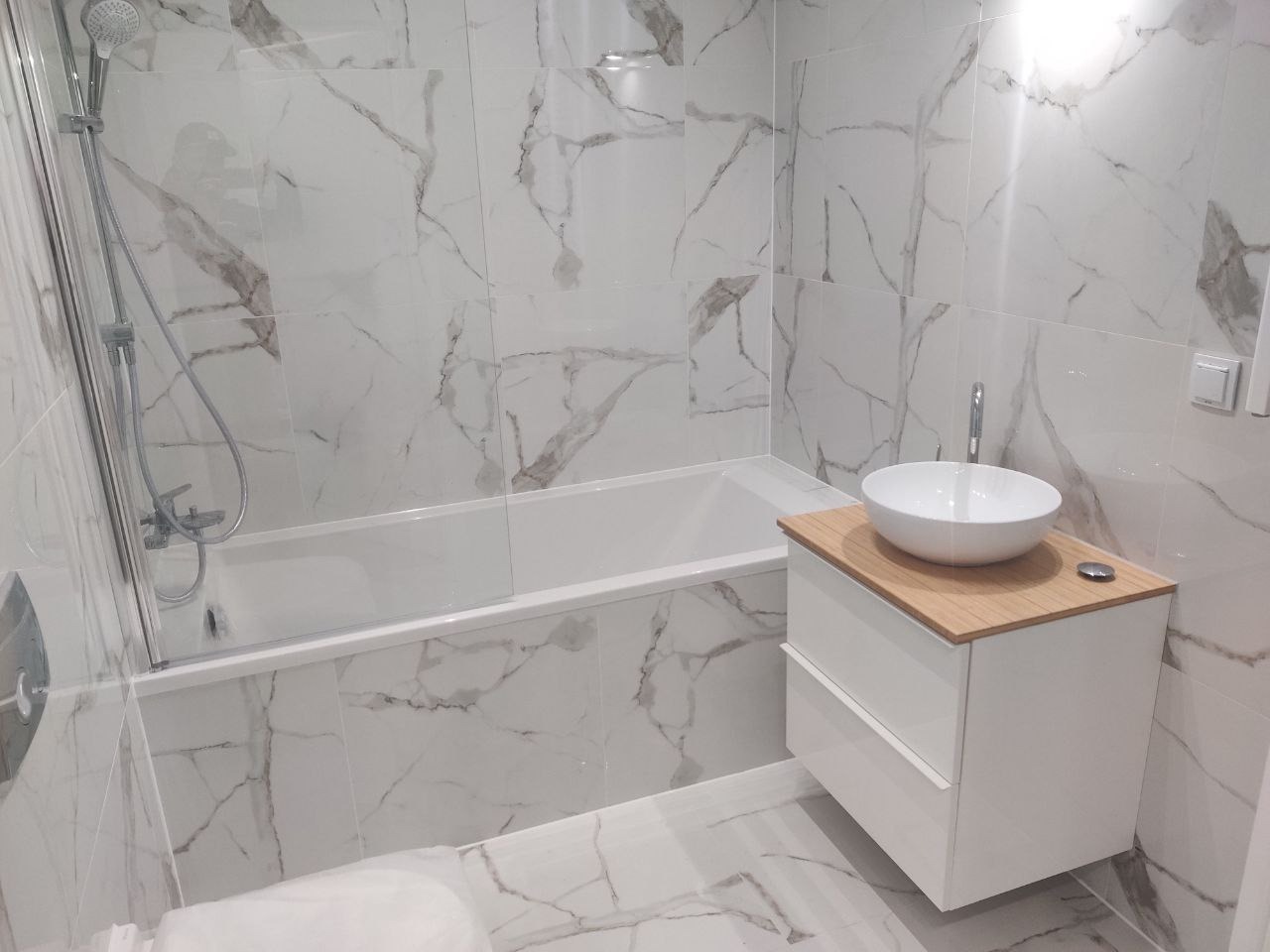 Wykończona łazienka z wanną obudowaną płytkami imitującymi marmur, białą szafką pod umywalką z drewnianym blatem i chromowaną armaturą.