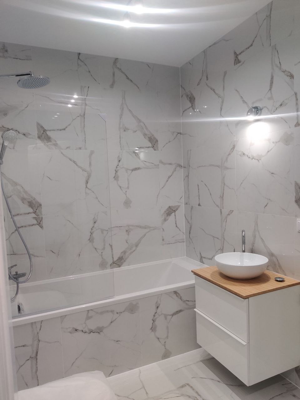 Biała łazienka z wanną i umywalką nablatową. Płytki imitujące marmur zdobią ściany. Nowoczesny design i jasne oświetlenie.