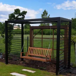 Metalowa konstrukcja z drewnianą huśtawką w ogrodzie, widok na błękitne niebo z chmurami. Minimalistyczny design, czarne metalowe elementy i naturalne drewno.