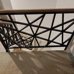 Arkadiusz Blat - Nowoczesna balustrada schodowa z czarnych, geometrycznych elementów i drewnianym poręczą, widziana z góry, oświetlona światłem padającym z góry.