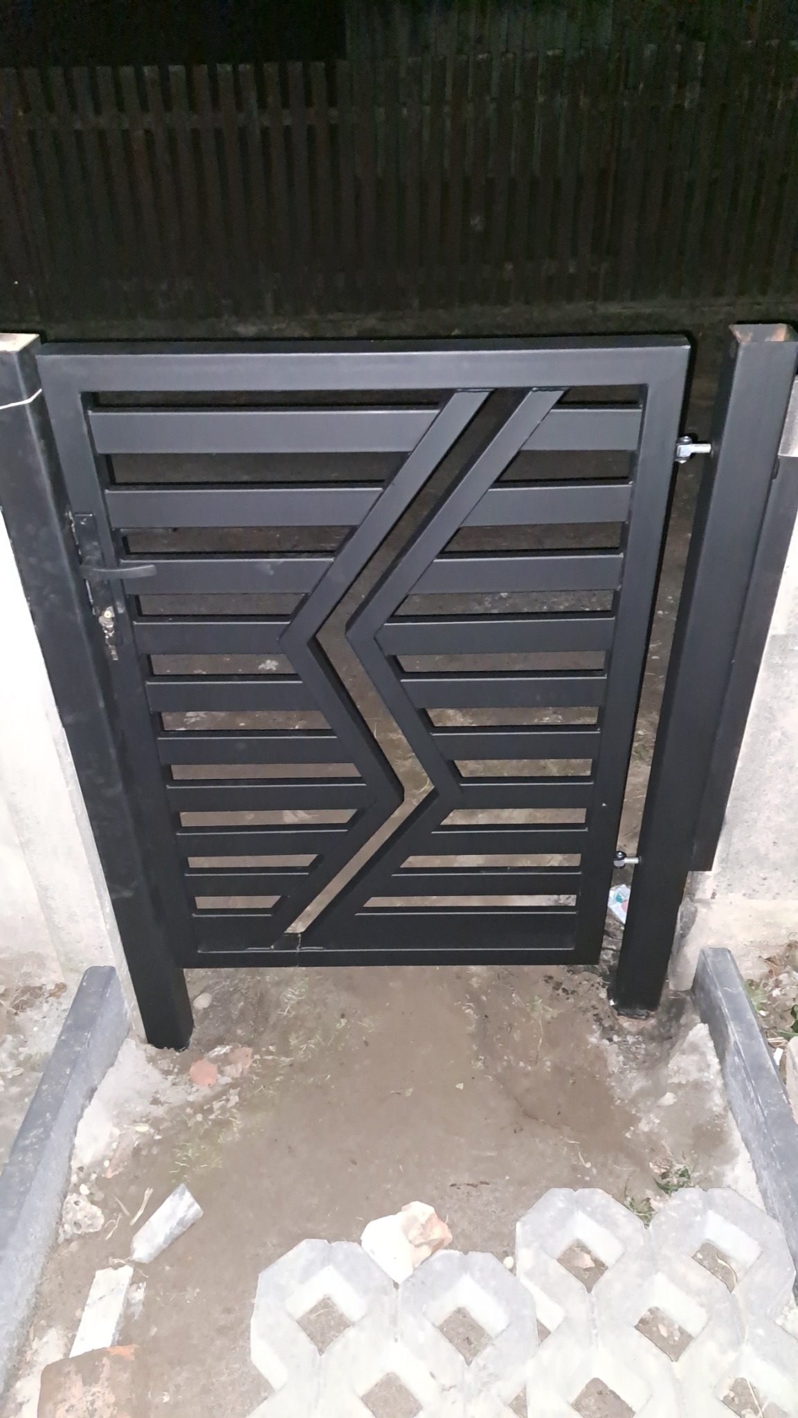 Nowoczesna, czarna furtka metalowa z poziomymi szczeblami i geometrycznym wzorem błyskawicy, zamontowana przy ogrodzeniu z drewnianych sztachet.