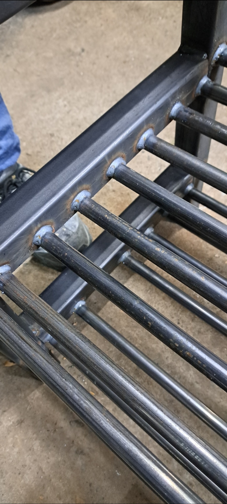 Metalowa konstrukcja z przyspawanymi prętami, widoczne spawy. Fragment ubrania i buta w lewym rogu kadru. Szlifowany metal.