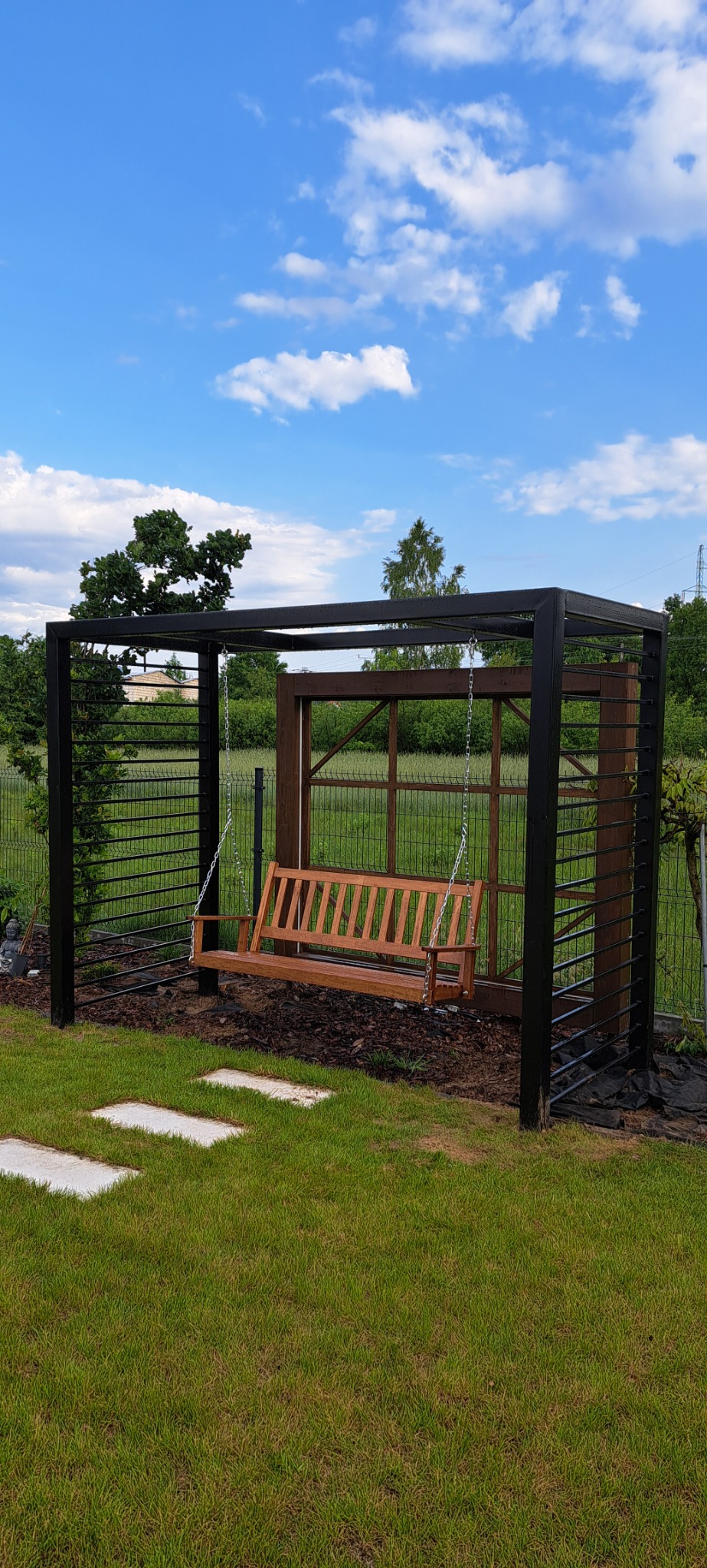 Metalowa konstrukcja z drewnianą huśtawką w ogrodzie, widok na błękitne niebo z chmurami. Minimalistyczny design, czarne metalowe elementy i naturalne drewno.