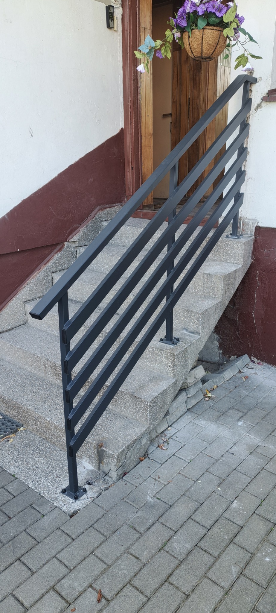 Zewnętrzne schody z szarą, metalową poręczą o minimalistycznym designie. Na górze wisi kosz z fioletowymi kwiatami. Widoczne szare płytki chodnikowe.
