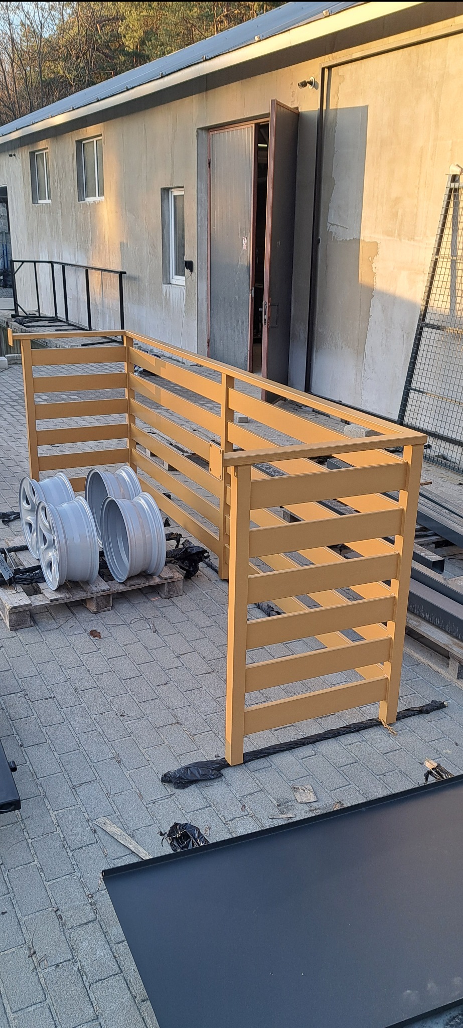 Fragment konstrukcji ogrodzenia z poziomych belek w kolorze piaskowym, oparta o paletę z felgami samochodowymi, na tle budynku gospodarczego.