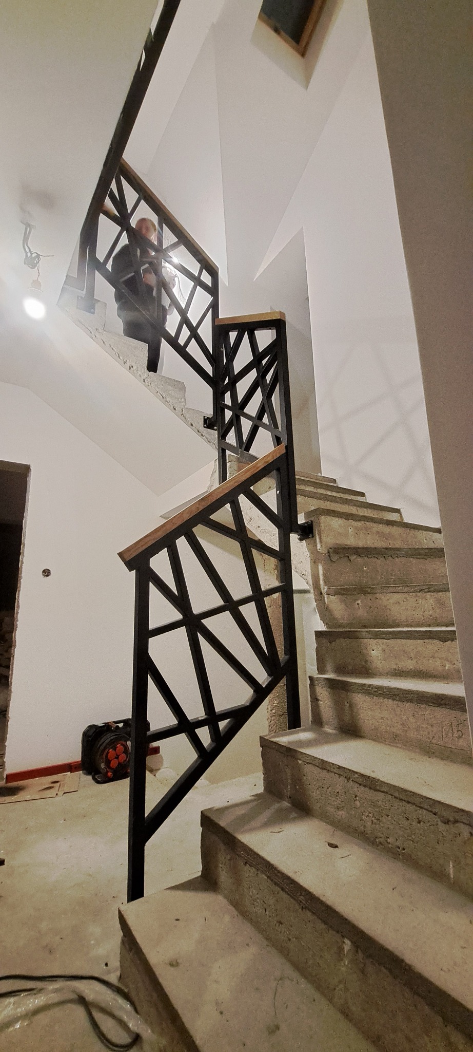 Nowoczesna, czarna balustrada schodowa z drewnianym poręczą, o geometrycznym wzorze, zamontowana na surowych, betonowych schodach w trakcie wykończenia.