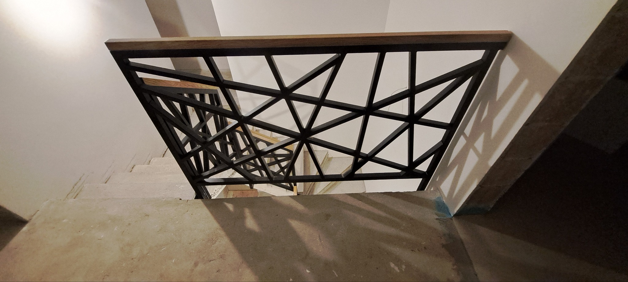 Nowoczesna balustrada schodowa z czarnych, geometrycznych elementów i drewnianym poręczą, widziana z góry, oświetlona światłem padającym z góry.