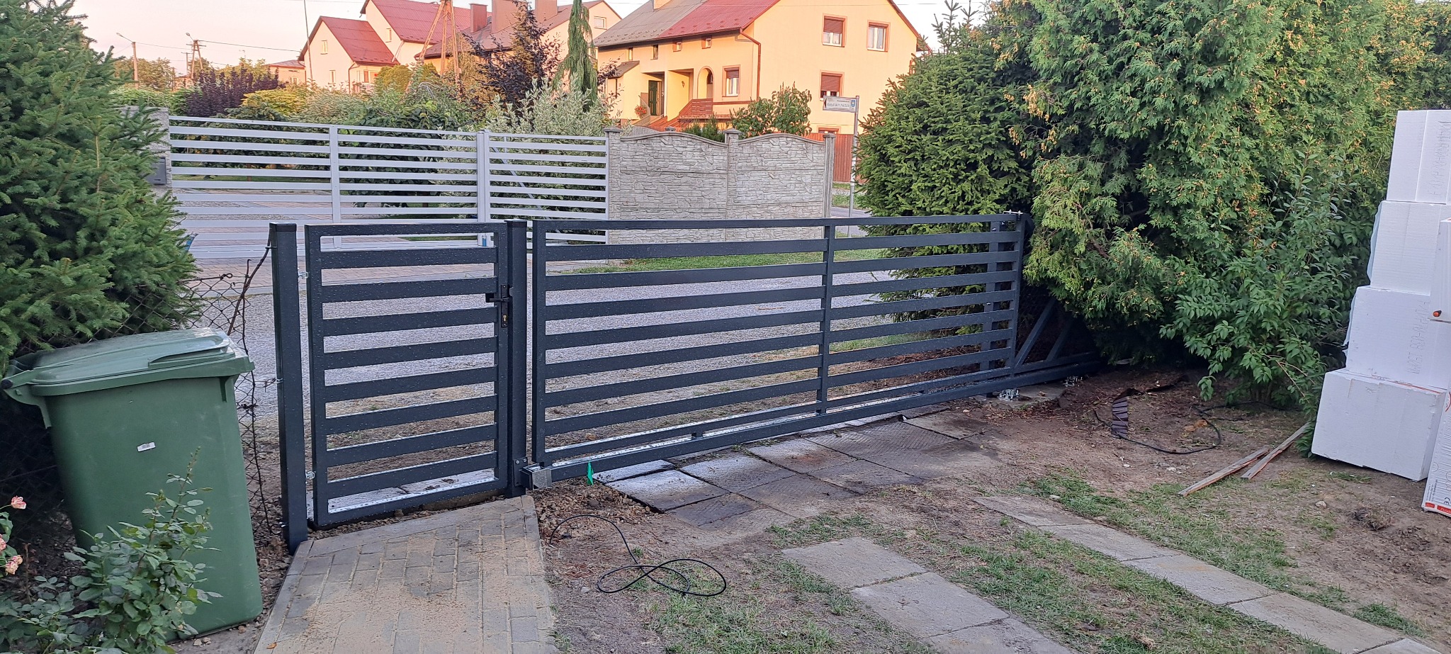 Nowoczesna, pozioma brama wjazdowa z metalu w kolorze grafitowym, z widocznymi elementami automatyki, na tle zieleni i zabudowań mieszkalnych.